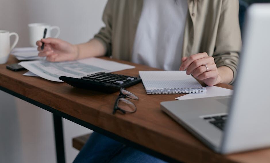 A woman manages finances at home, using a laptop and calculator on a wooden desk.
