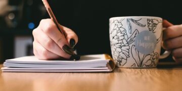 Person writing in a notebook with a floral ceramic mug on a wooden desk.